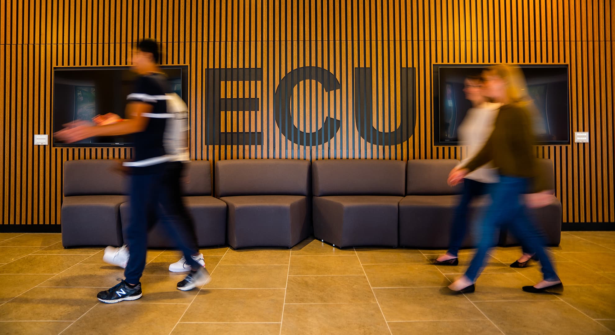 students walking past ECu sign on intricate wooden wall.