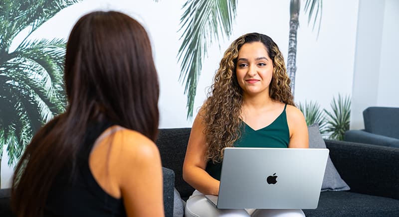 two women chatting on a couch, one with a laptop