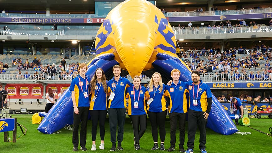 Female and male students on an Australian Rules football ground in front of a crowd.