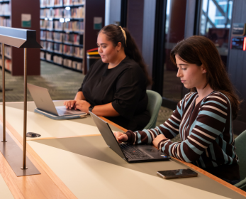 Two students using laptops at a study desk inside the ECU Library, with bookshelves and study spaces visible in the background. One student is seated at a larger workstation while the other works at an individual desk, highlighting quiet study and digital learning spaces within the library.