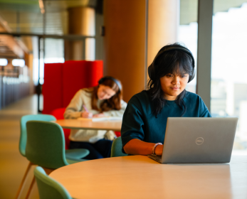 Student using a laptop to access online library resources while studying in a modern university library space, with other students working in the background.
