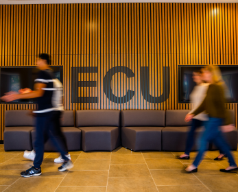 Students walk through the ECU South West Campus, passing the ECU feature wall and seating area. Motion blur captures movement through the space, conveying a welcoming and active campus environment.