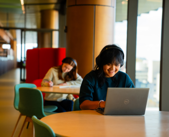 Student using a laptop in the ECU City Campus Library, with another student studying nearby and large windows overlooking the city.