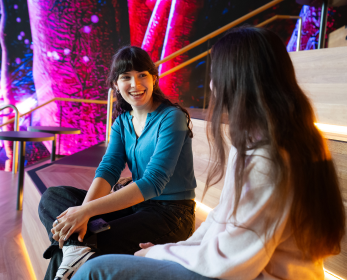 Two students sitting together at ECU City Campus, smiling and chatting on tiered seating with colourful digital artwork in the background.