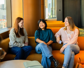 Three students sit together on a curved lounge in a bright library space, smiling and chatting. Large windows and warm timber finishes create an inviting atmosphere that reflects connection, belonging and community.