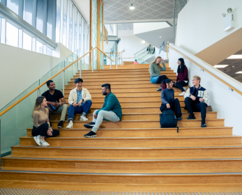 Students seated on a large timber staircase inside the ECU Joondalup Campus Library. Some are chatting in small groups while others are sitting individually, with large windows filling the space with natural light.