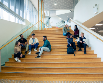 A group of students sit and talk on wide timber steps inside the Joondalup Campus Library, with large windows and an upper staircase in the background.