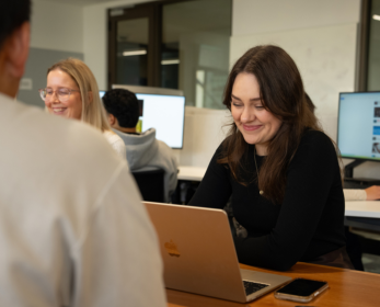 A woman smiling while using a laptop at a table in a computer lab, with other people working at computers in the background.