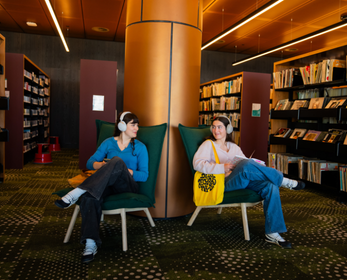 Two students wearing headphones sit facing each other in lounge chairs inside the Library, smiling as they chat. One student is carrying a bright yellow tote bag, with bookshelves and warm timber finishes visible in the background.