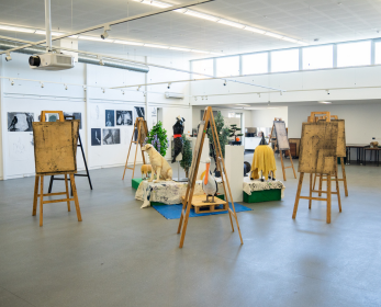 An open art studio with several wooden easels displaying artworks arranged throughout a bright, spacious room. Additional artworks are displayed on tables and hung along the walls, with large windows providing natural light.