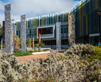 Exterior of ECU Joondalup Campus Library, featuring a modern glass façade with colourful vertical panels, decorative metal sculptures and native Australian landscaping in the foreground.