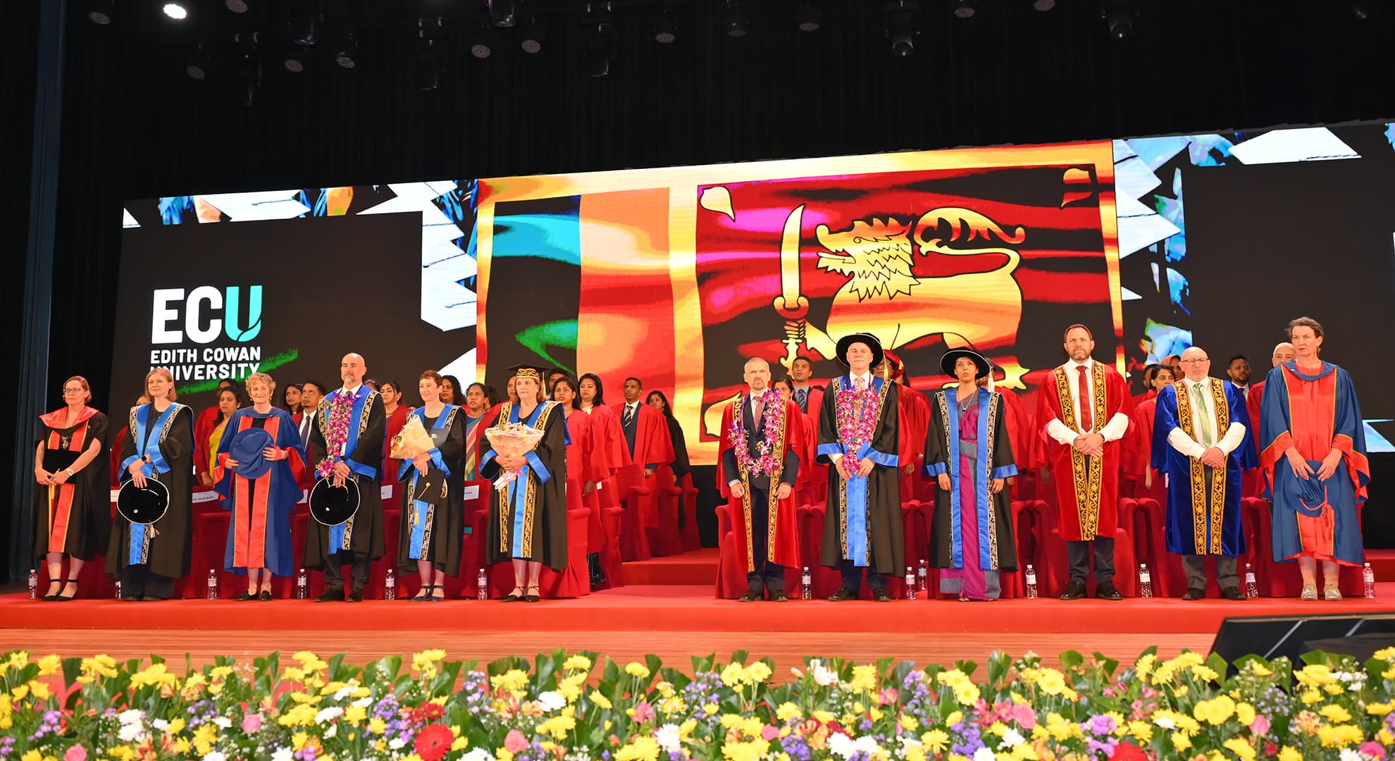 People in graduation regalia standing in front of a screen.