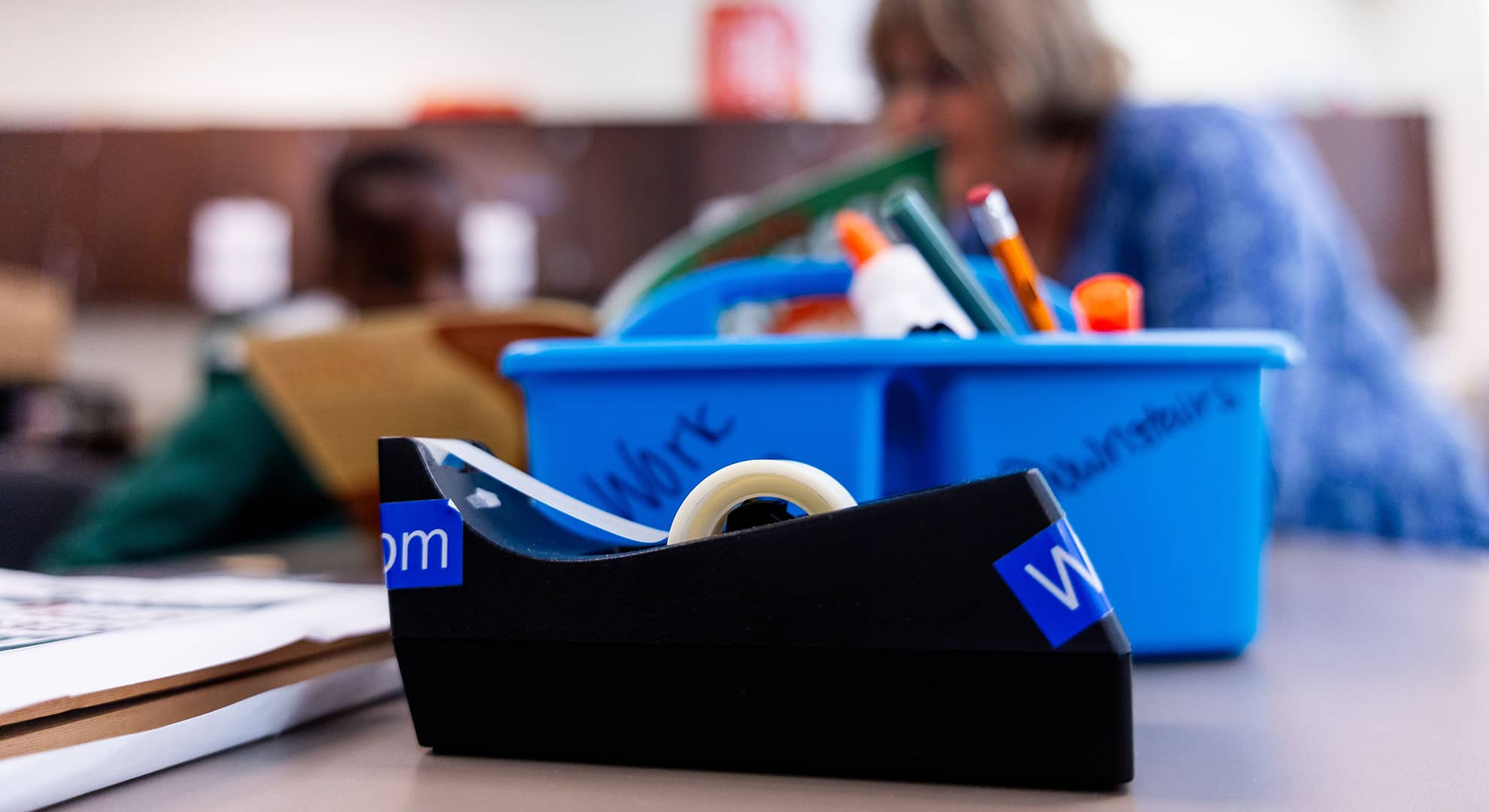 Close up of tape dispenser, with teacher and student out of focus in the background.