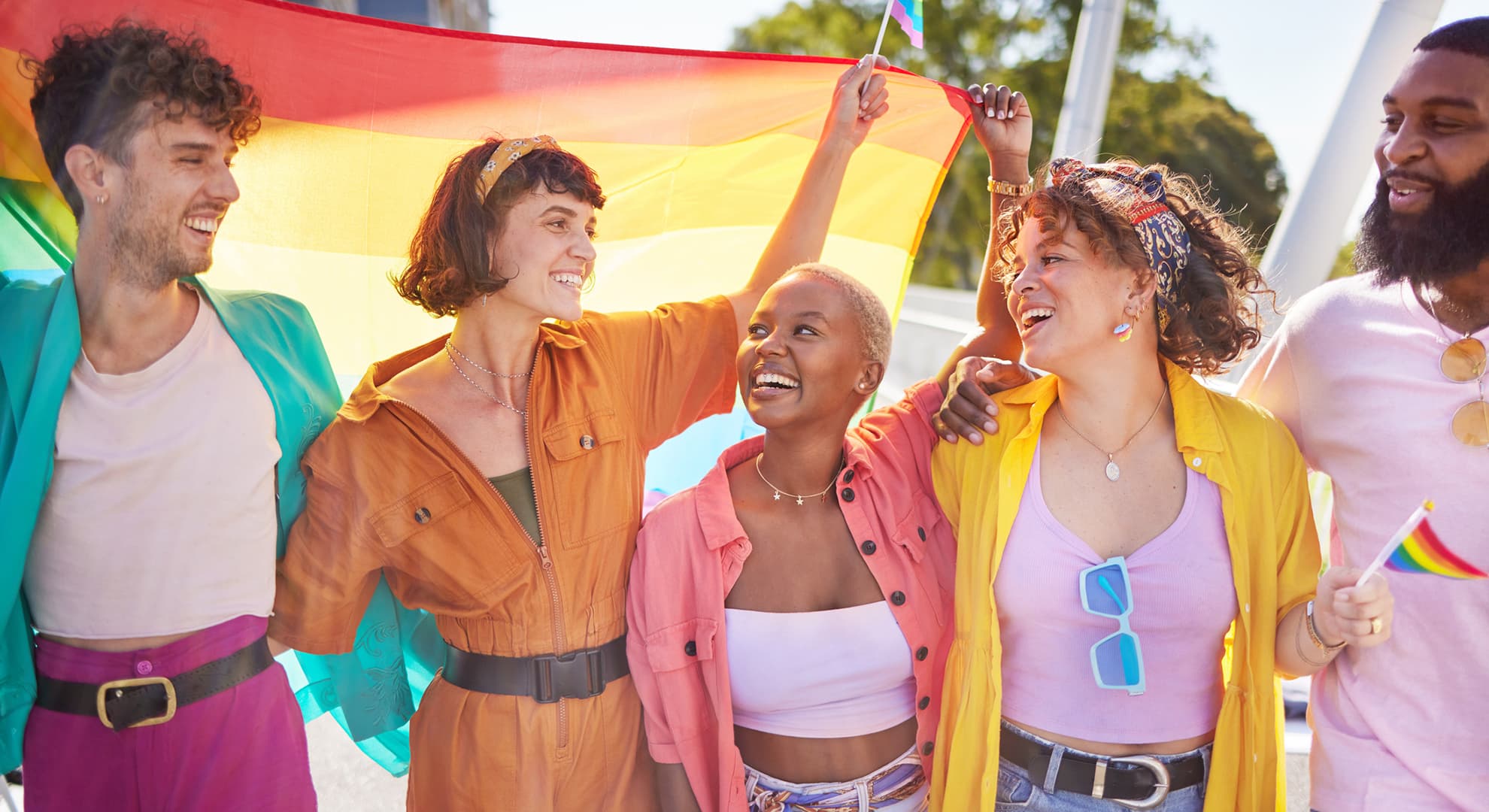 Five people in colourful clothes standing under a pride flag while smiling.