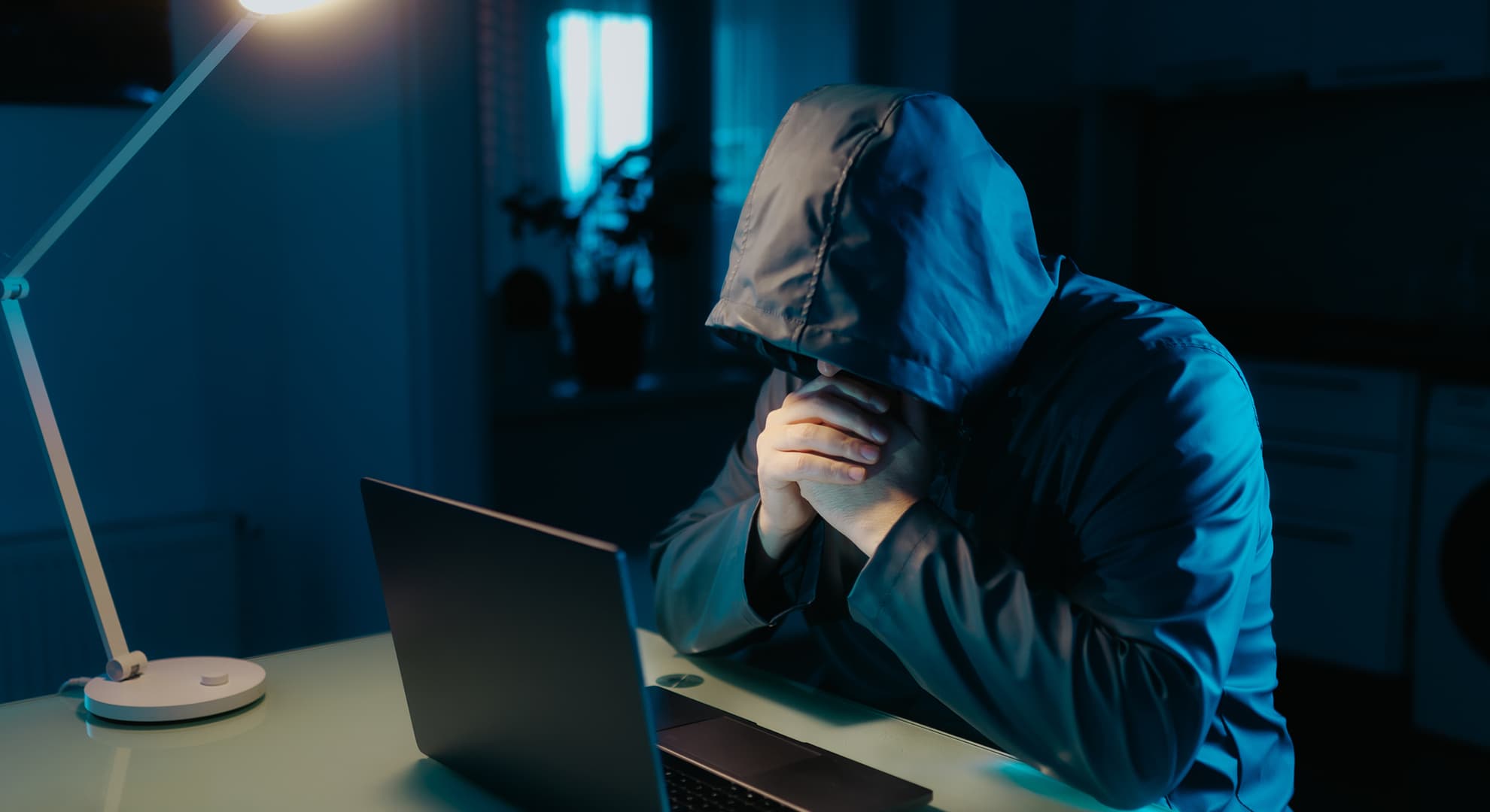 Man with hooded jumper sitting in dark room at computer.
