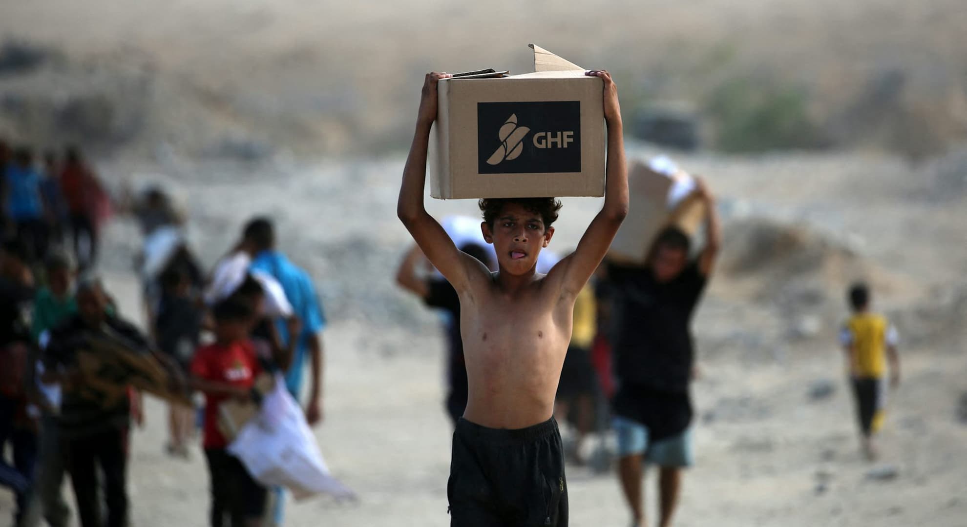 Shirtless Palestinian boy walking with GHF humanitarian aid box on his head.