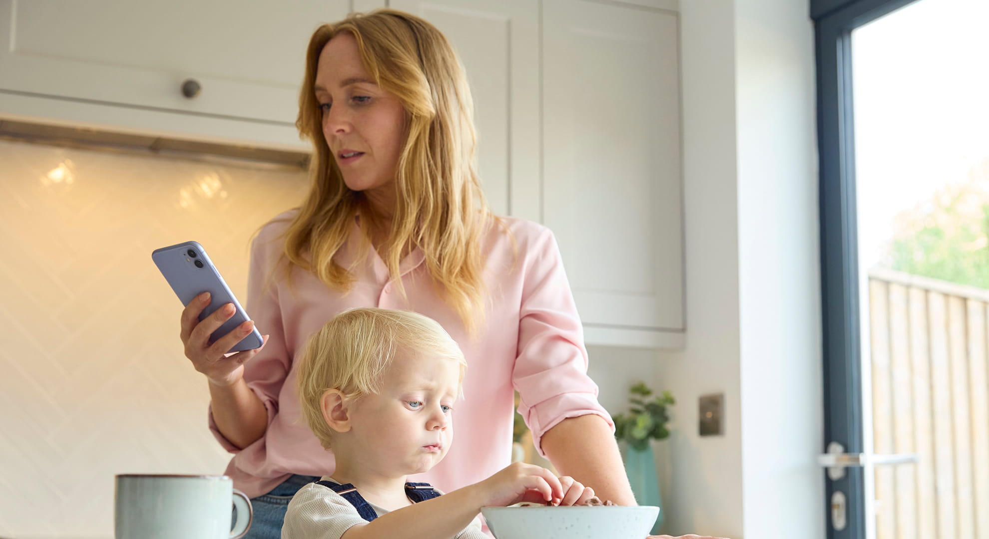 Mother at home looking at mobile phone as young son eats breakfast in kitchen.