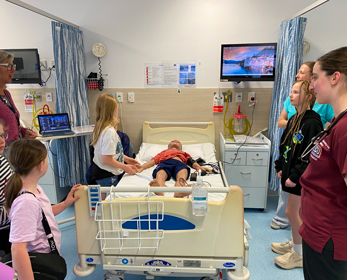 Children participate in a Children&rsquo;s University school holiday workshop in a simulated hospital ward. A group gathers around a hospital bed where a medical training mannequin lies connected to monitoring equipment. University staff and nursing students demonstrate healthcare procedures while children observe and interact with the clinical learning environment. Medical equipment, bedside monitors and training resources are visible throughout the room.