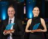 Two award recipients stand side by side at an indoor awards event, each holding a blue glass trophy mounted on a wooden base. The person on the left, Christopher Lambe, is wearing a dark suit, white shirt and patterned tie. The person on the right, Nicolette Beard, is wearing a sleeveless black outfit and statement earrings. The background features blue stage lighting and softly blurred event décor.