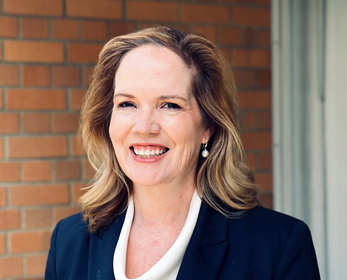 Professional headshot of a person wearing a dark blazer over a light-coloured blouse, standing in front of a red brick wall and a window frame. The image is cropped from the chest up, with natural lighting and a softly blurred background. Sarah is facing the camera in an outdoor corridor setting.
