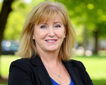 Professional headshot of a person with shoulder-length light-coloured hair, wearing a black blazer over a bright blue top and a silver necklace. The person is photographed outdoors, facing the camera, with a blurred background of green trees and parkland.