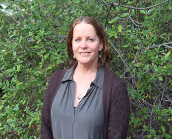 Ms Caroline Neske standing outdoors in front of dense green leafy shrubs, facing the camera. The person is wearing a charcoal-grey collared blouse and a dark knitted cardigan, with a pendant necklace visible. The image is a waist-up portrait taken in natural daylight, with foliage filling the background.