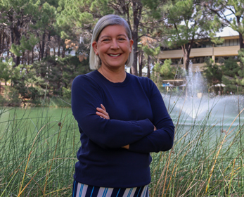 Professional headshot of Dr Michelle Striepe standing outdoors with arms crossed beside a lake, surrounded by reeds and trees, with a water fountain and campus-style buildings visible in the background, she is wearing a navy long-sleeved top and striped trousers.