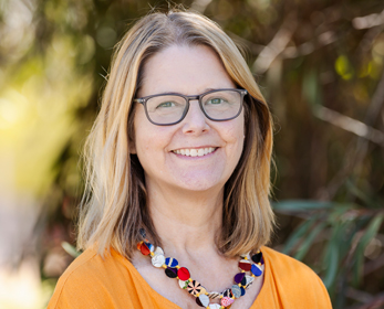 Professional head-and-shoulders portrait of Dr Susan Main with shoulder-length hair and glasses, wearing an orange top and a colourful beaded necklace, standing outdoors with blurred green foliage in the background.