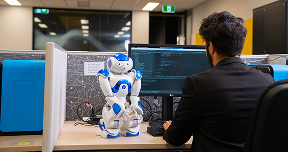 small robot on a desk and a man looking at a computer screen.